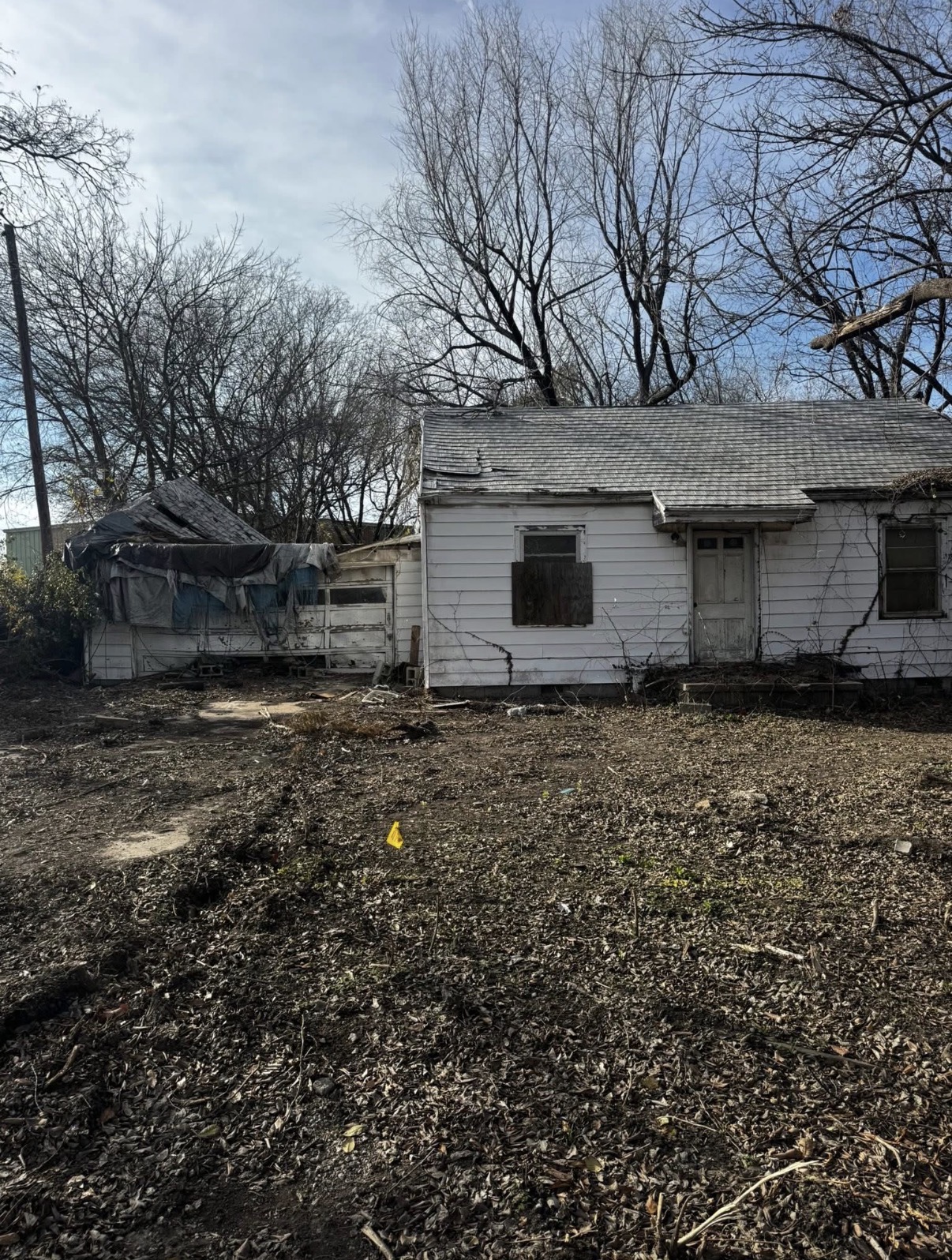 Abandoned white shack with tarps, boarded windows and overgrown debris before demolition